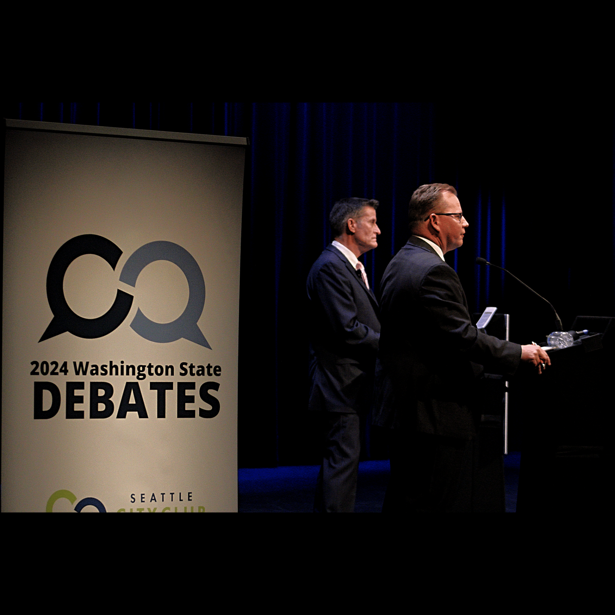 Chris Reykdal (Close) and David Olson (Far) standing at the podiums during the 2024 Superintendent of Public Instruction debate.